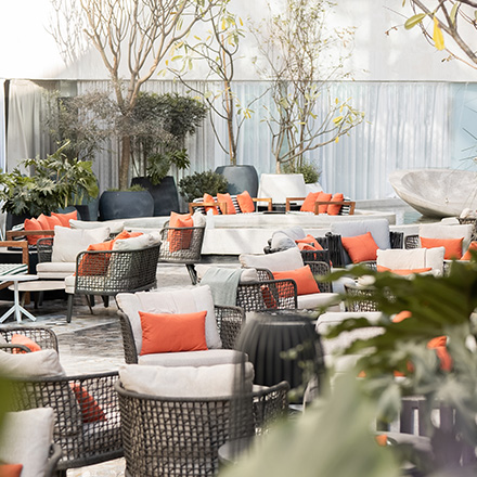 Outdoor terrace with cream cushioned outdoor lounge seats with orange pillows and plants and trees scattered throughout the space.