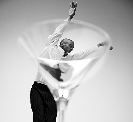 Black and white image of man in a white shirt and dark pants dancing with arms up in the background, looking through a clear martini glass in the foreground.