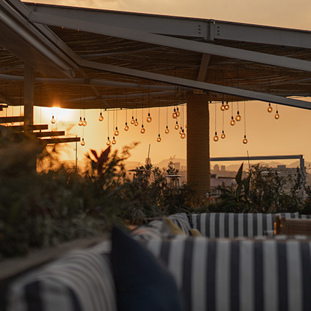 Terraza al aire libre con la puesta de sol al fondo, asientos de cojines a rayas en primer plano y techo del establecimiento.