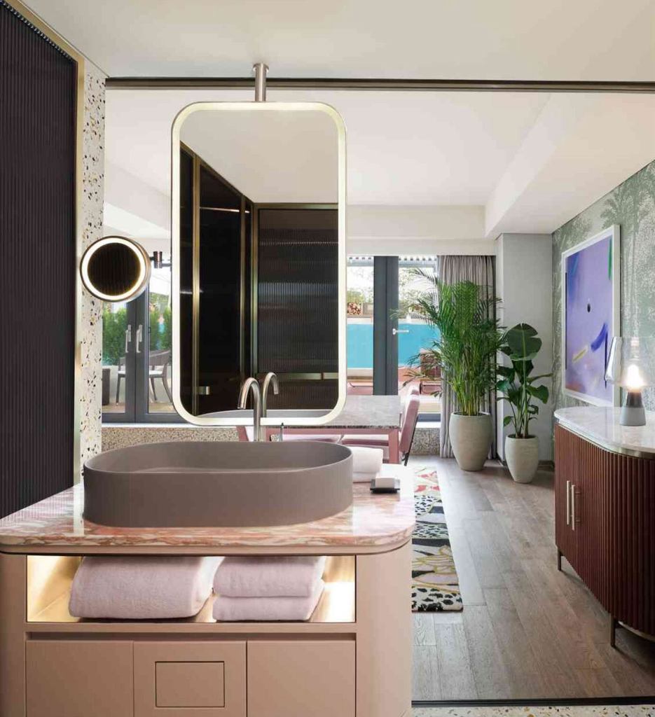 Pink marble top bathroom sink with grey bowl and modern lit up framed mirror with white towels folded underneath the sink, a living room is outside the glass walls of the bathroom