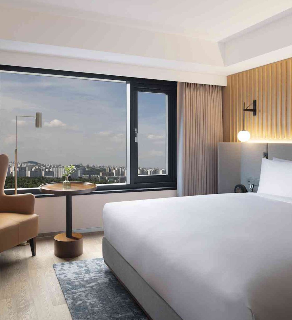 Large white bed with grey headboard on grey pattern rug with tan chair in the corner with a small round table and windows overlooking the city