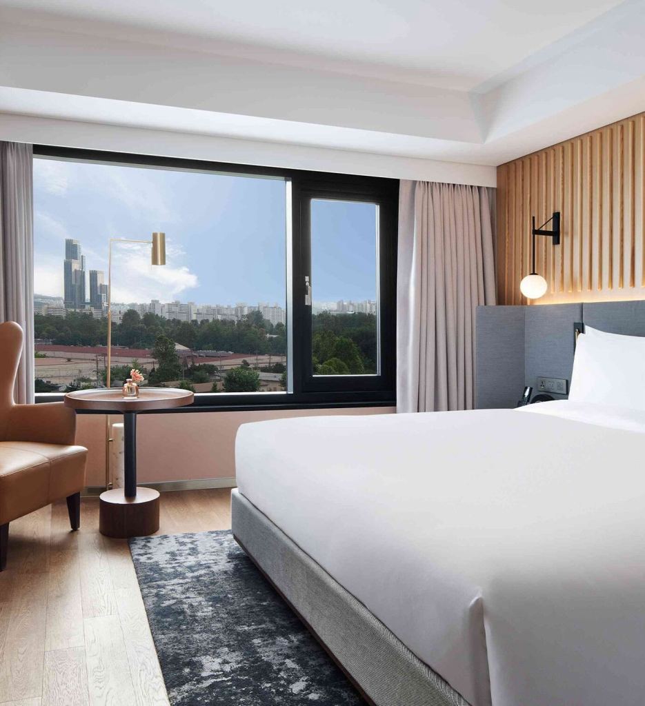 Large white bed with grey headboard on a dark pattern rug with a tan leather chair and bronze small round table in the corner and windows overlooking a city