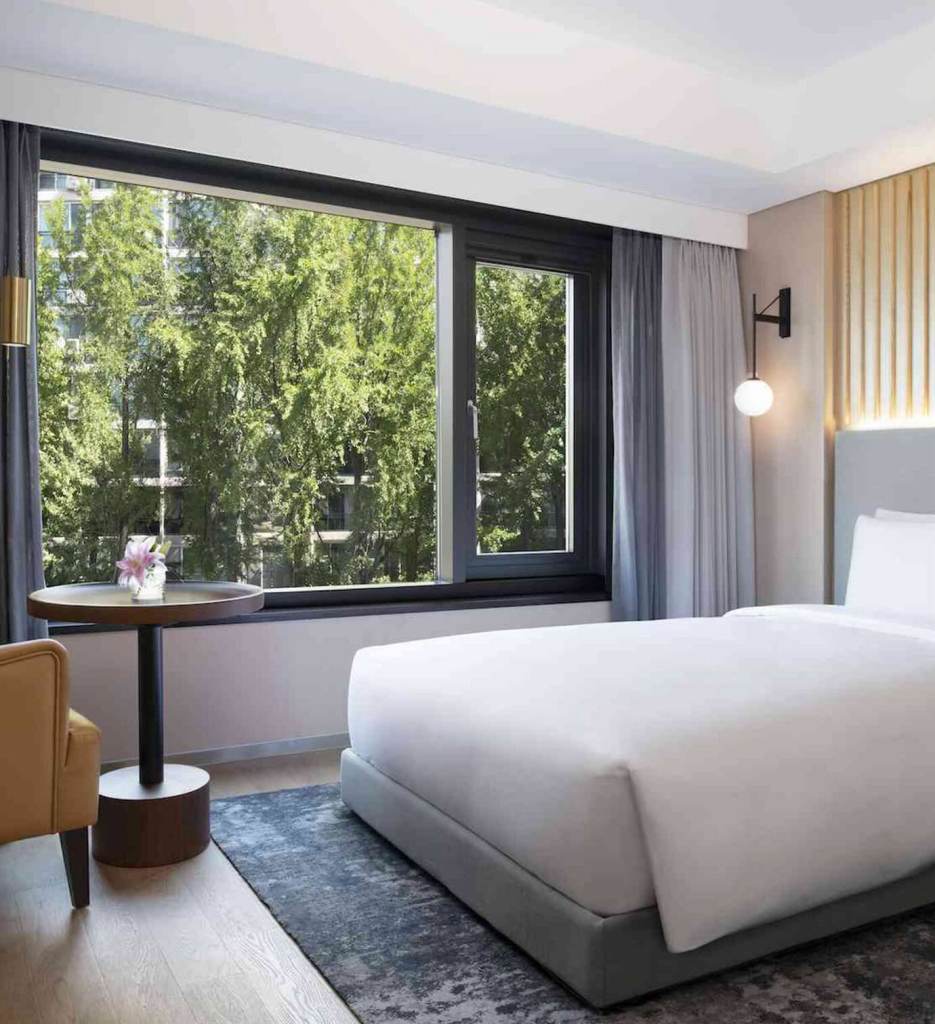 Single bed with light grey headboard on a pattern rug with the corner of a tan chair in the corner with a small round table and a window overlooking trees