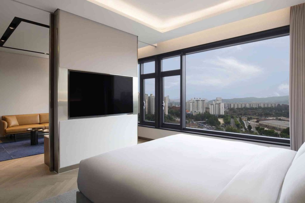 Large room with white bed, plasma screen tv, tan sofa in the room over, and a large window overlooking the city