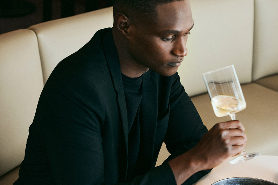 A man in a black suit jacket and t-shirt drinking a glass of white wine.