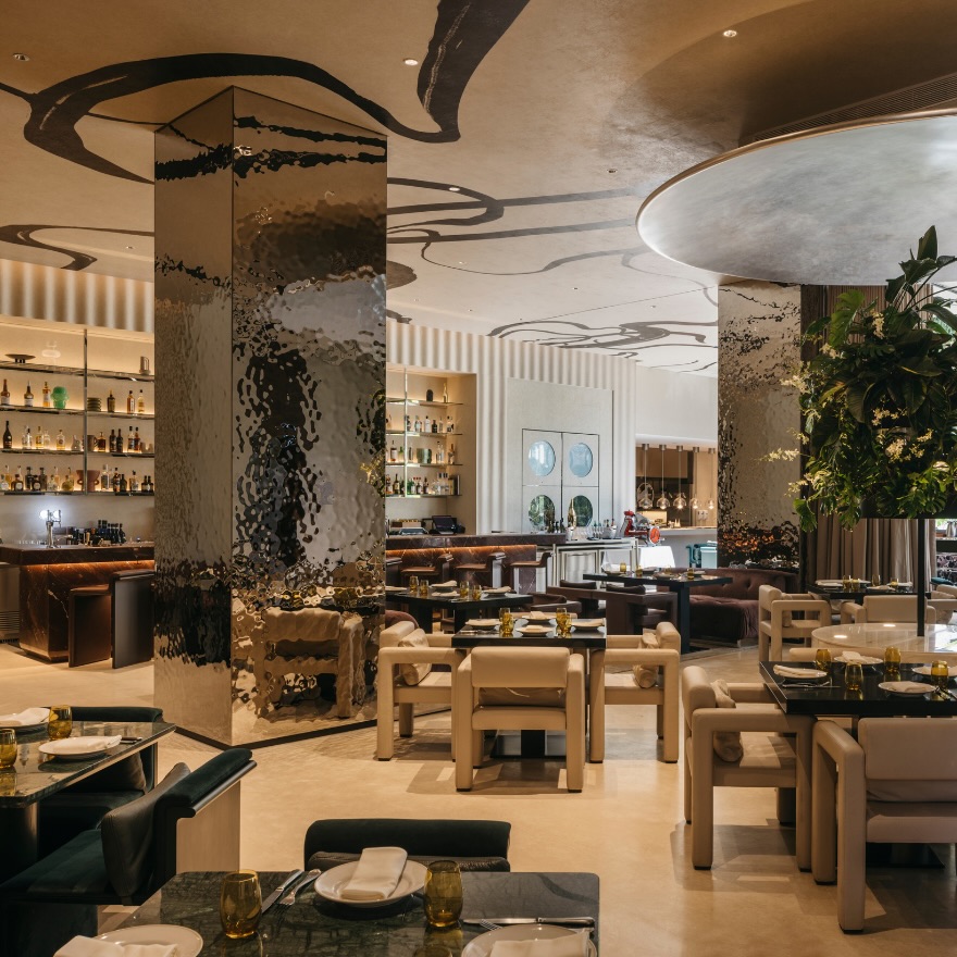 A dining room in a restaurant with a tree in the right, a bar in the distance, modern, neutral-hued sculptural seating and a large square column with a reflective finish