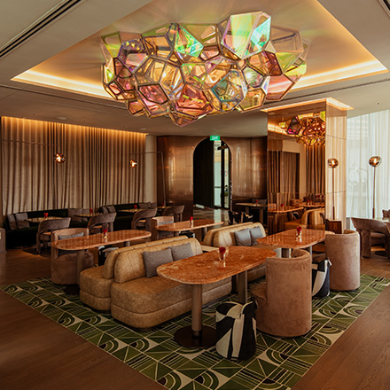 Warmly lit lounge space with geometric chandelier, green patterned area rug, and light brown cushy booth seating with oval tables.