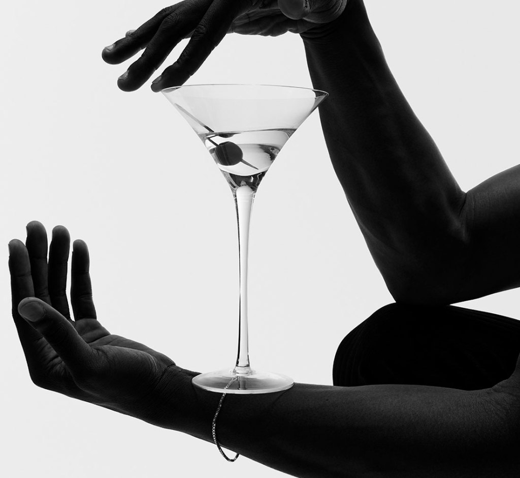 Black and white image of hands framing a clear martini glass with an olive floating inside.