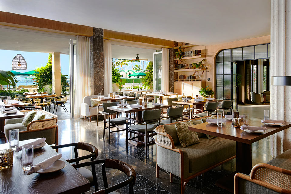 Indoor-outdoor dining room with rows of square and rectangular wooden tables with various benches and chairs. Two large doors look out to an outdoor terrace filled with green plants.