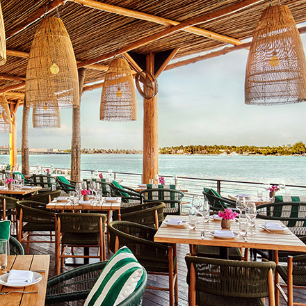 Patio al aire libre con luces colgantes de mimbre, techo de mimbre, pequeñas mesas de madera y sillas de estilo exterior con vistas a un mar azul.