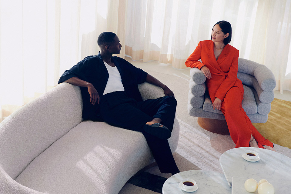 A man dressed in a black two piece outfit, and a woman in an orange jumpsuit, sitting in chairs enjoying a cup of coffee.