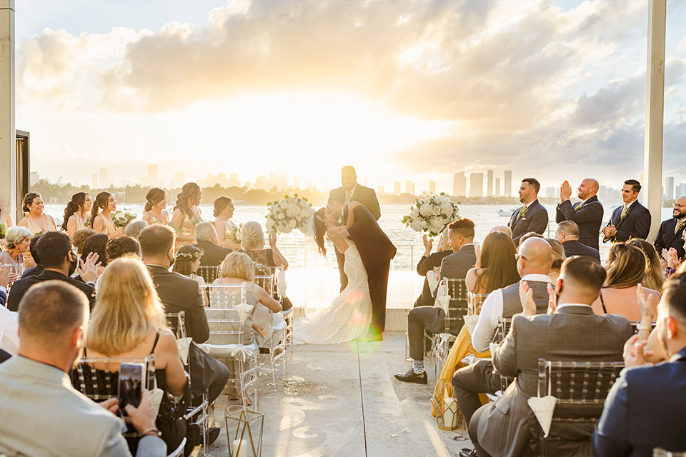 Ceremonia de boda con vistas a la bahía durante el atardecer, con una novia y un novio besándose mientras la fiesta nupcial, los invitados y el oficiante aplauden.