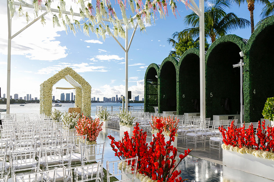 Disposición de una ceremonia de boda con vistas a la bahía, sillas transparentes y flores blancas y rojas que bordean el pasillo.