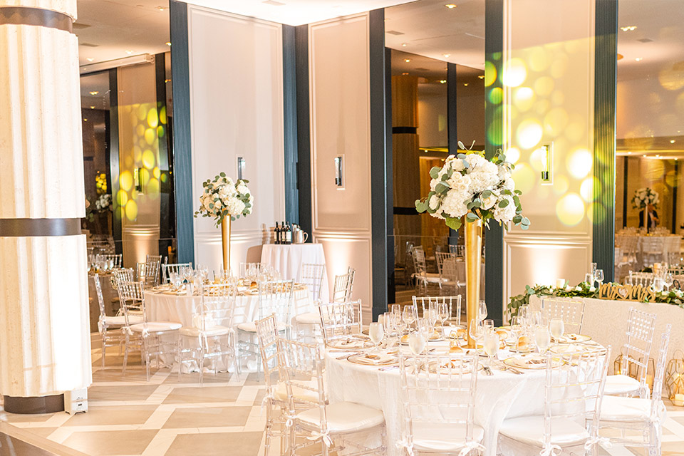 Gran salón preparado para una boda con mesas redondas, sillas transparentes y arreglos florales elegantes en jarrones altos dorados.