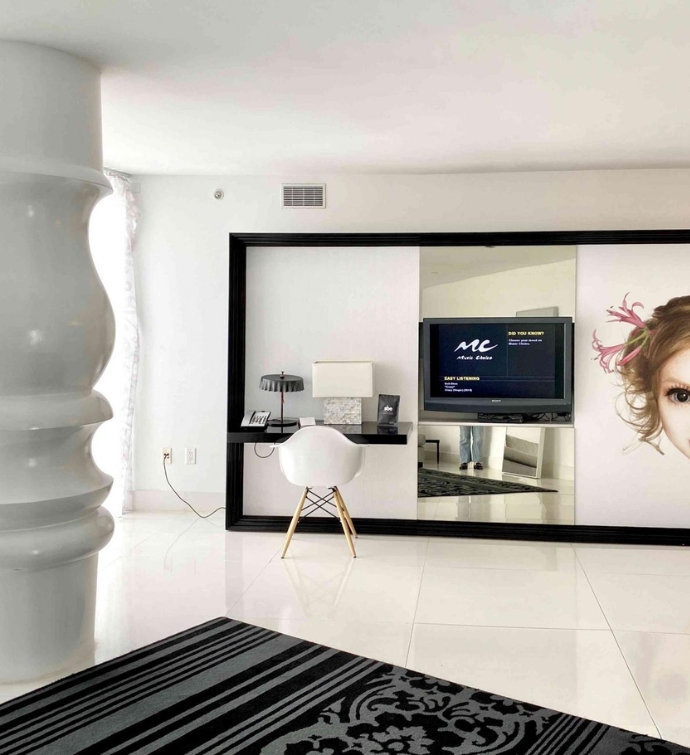 Large white room with white desk chair, black desk, and plasma screen tv with large white column in the middle of the room and a patterned rug