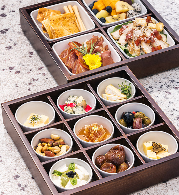 a box with 9 sections, each featuring a bowl with a different small snack, near a second box with 4 sections of small plates