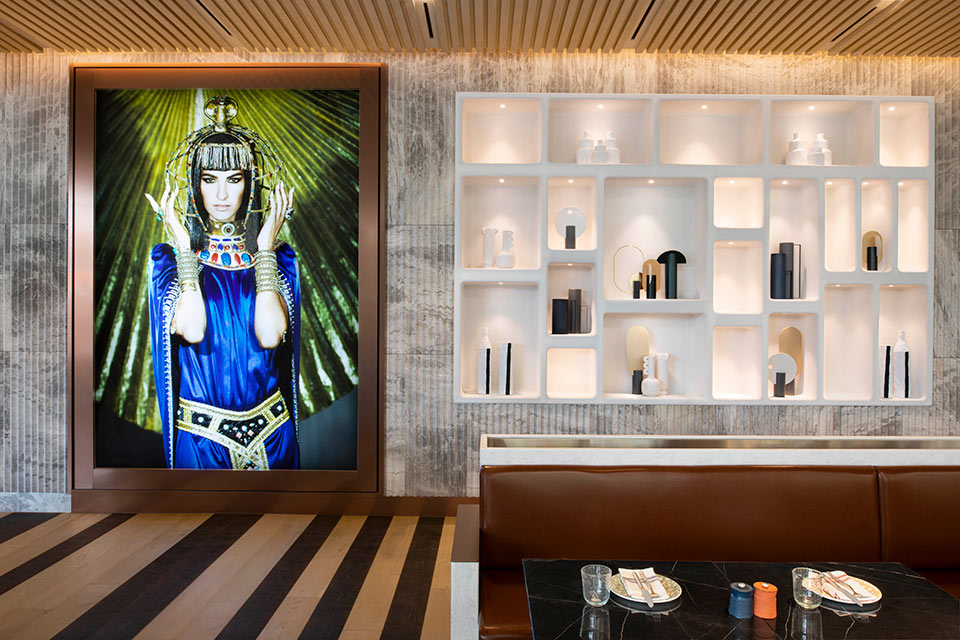 A vibrant floor to ceiling photograph of a woman in blue draped dress and gold jewels hangs on the wall alongside white shelving showcasing objet at the restaurant Cleo Seoul