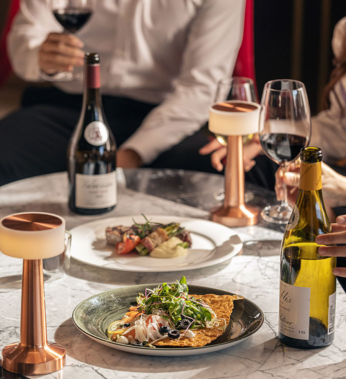 Low table with 2 dishes and wine glasses, and a man in the background holding a glass of wine.