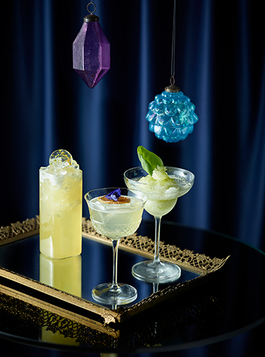 Three yellow cocktails in different style glasses sitting on a mirrored tray underneath two hanging crystals