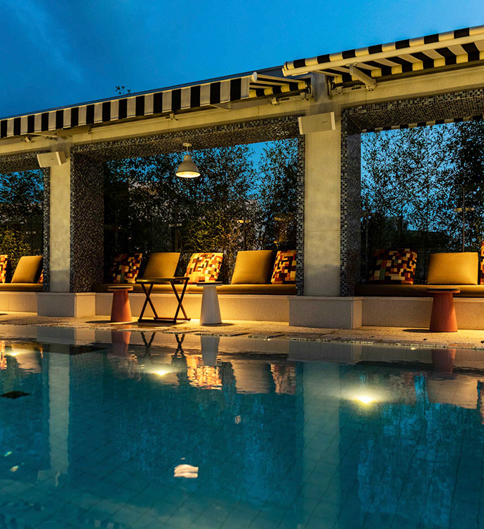 An outdoor swimming pool lit up at night with a black and white striped awning over poolside seating in the background