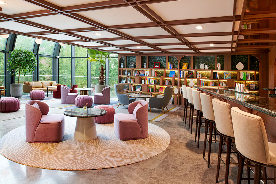 Lounge space with cream circular textured rug with pink velvet lounge chairs, circular mushroom coffee table, and more seating in the background with wooden shelves.