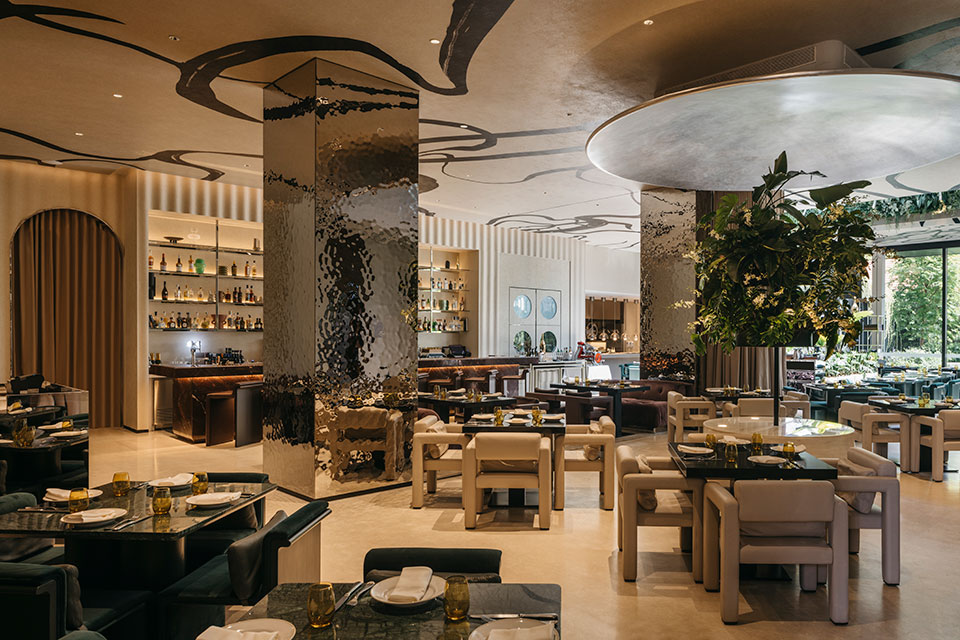 A dining room in a restaurant with a tree in the right, a bar in the distance, modern, neutral-hued sculptural seating and a large square column with a reflective finish