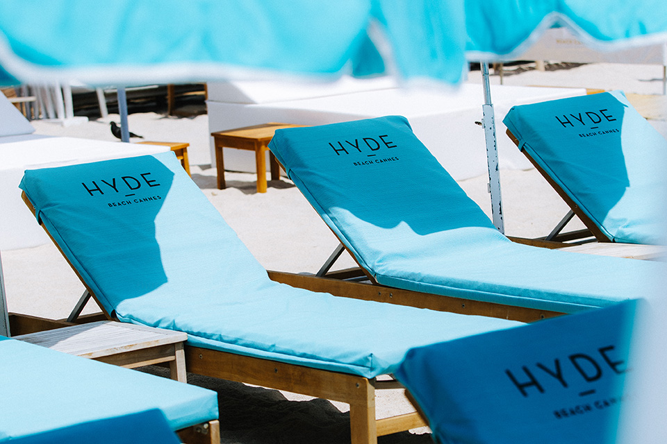 Light blue lounge chairs, with the Hyde Beach logo on the chairs, lined up on the beach under light blue umbrellas 