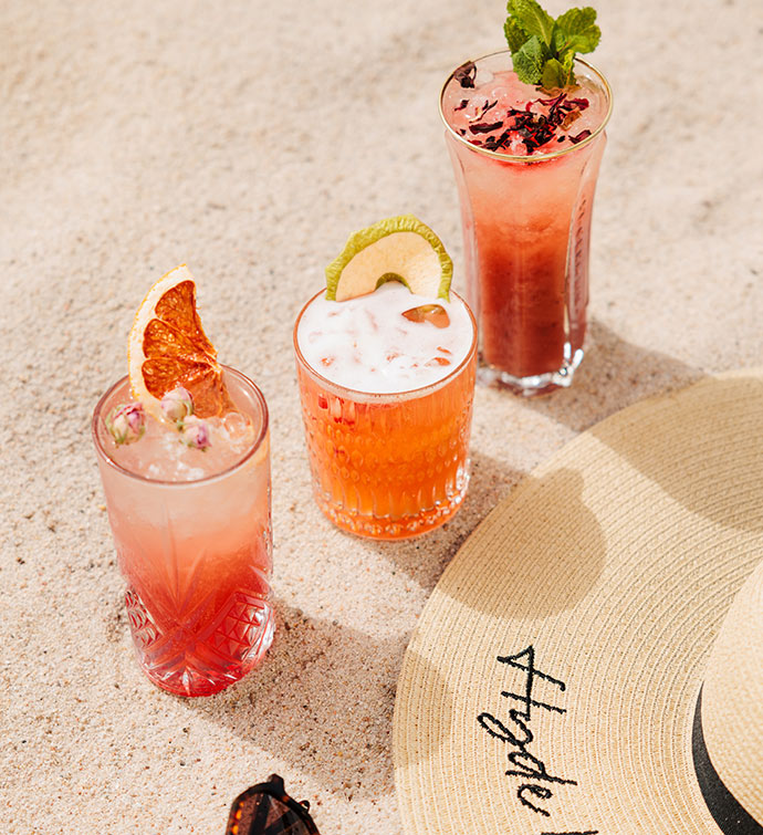 Trois cocktails aux teintes rouges à côté d'un chapeau de soleil portant une broderie Hyde