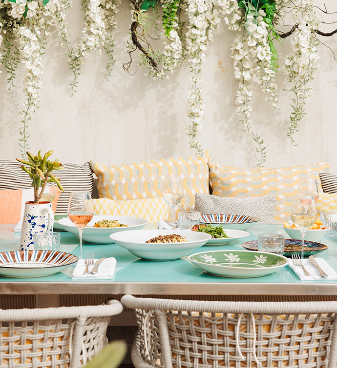 Une table d'extérieur avec des chaises en rotin tressé et un banc garni de coussins, photographiée en journée, sur laquelle est dressé un repas accompagné de rosé et où se trouvent des sièges sous des fleurs blanches