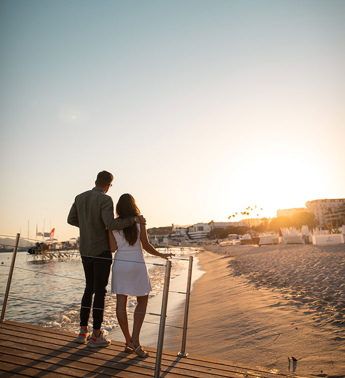 Un hombre y una mujer fotografiados de espaldas, de pie en la playa y contemplando la puesta de sol