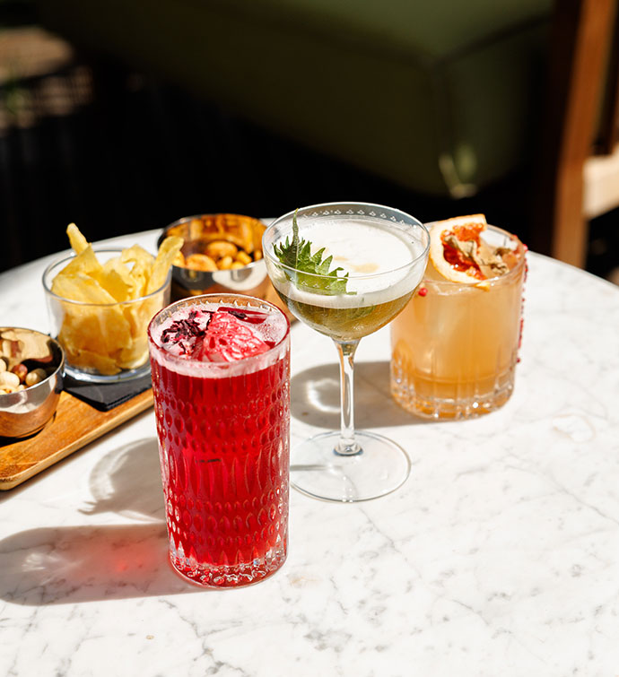 Three colorful cocktails alongside bar snacks on a marble table