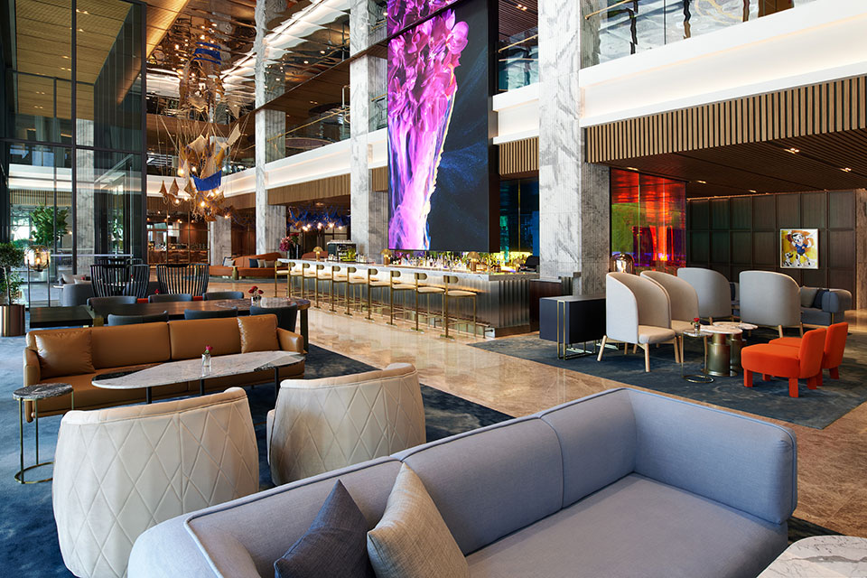 Sleek lounge space with light grey sofa in the foreground and cream lounge chairs and modern bar in the background with golden design accents throughout the space.