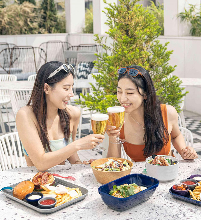 Two women making a toast with beer, over a spread of food being enjoyed on an outdoor patio