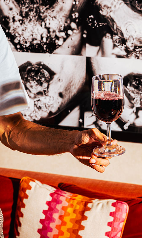 An arm holding out a glass of red wine, with bold black and white artwork hanging on the wall over a brightly colored and patterned pillow