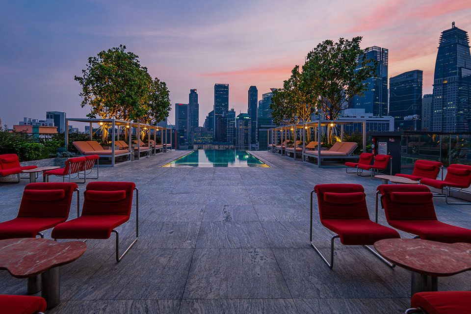 Rooftop pool, at dusk, with ruby red lounge chairs in the foreground and the Singapore skyline in the distance