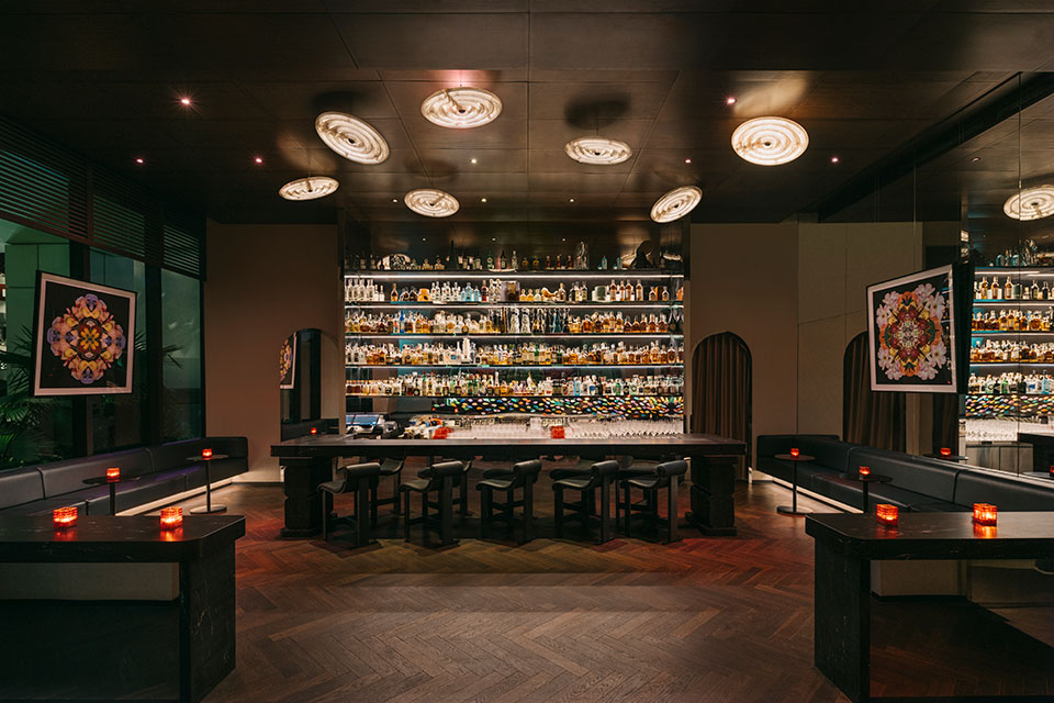 Bar with bottles on backlit shelves to the ceiling, in front of black barstools, and black banquets lining the room