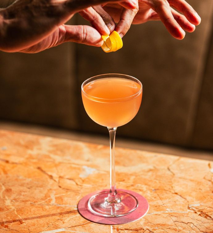 Orange cocktail in a tall, slender coup glass with lemon garnish being placed