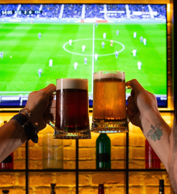 Dos manos brindando con cerveza con un partido de fútbol en la televisión