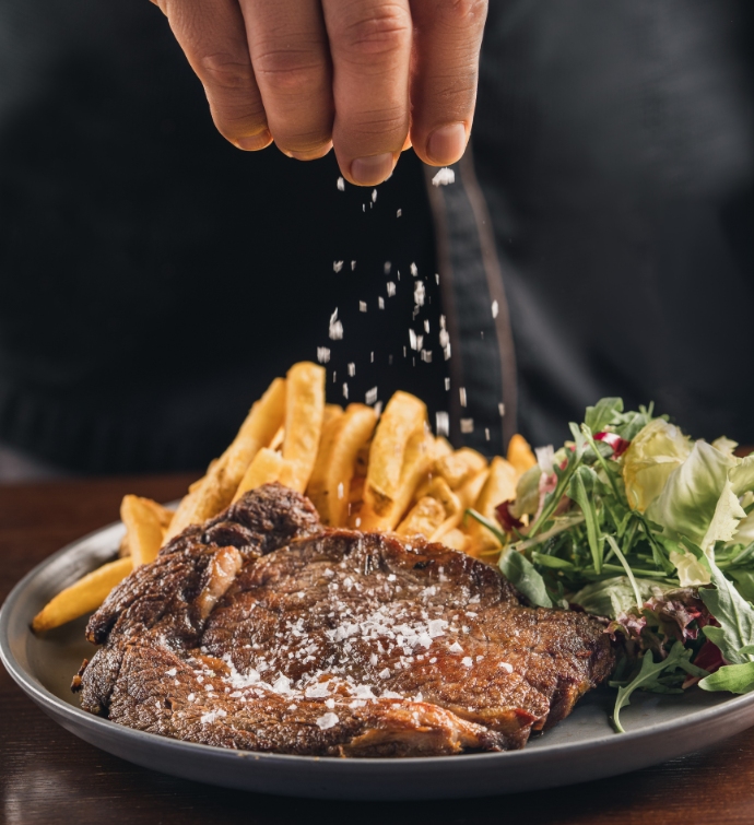 Mano poniendo sal en un filete con patatas fritas y ensalada de guarnición