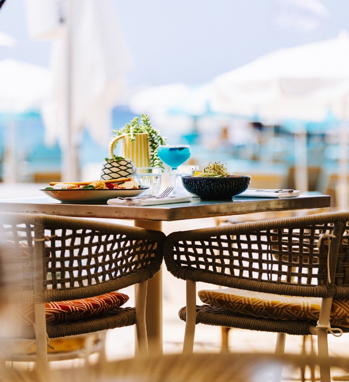 mesa de comedor al aire libre con cócteles y platos de comida y sombrillas al fondo