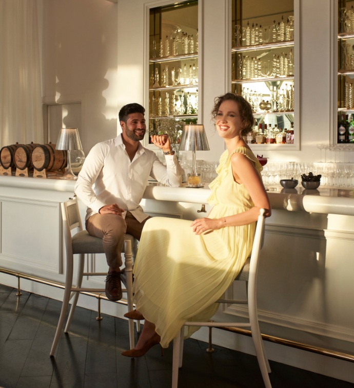 Femme vêtue d'une robe jaune et homme en chemise blanche et pantalon beige assis au bar