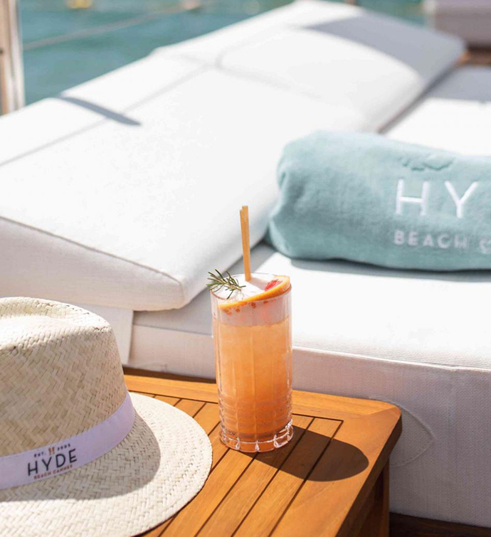 Close up image of a white chaise lounge bed with light blue rolled up towel laying on it and a brown side table with orange cocktail and small straw hat