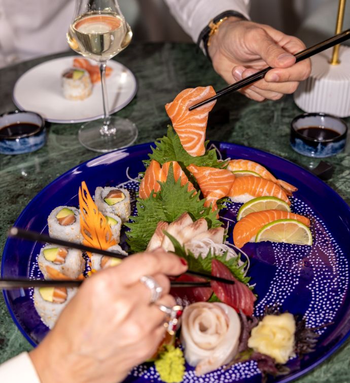 Sushis et sashimis sur une assiette bleue avec un verre de vin en arrière-plan et deux mains tenant des baguettes pour ramasser des sushis