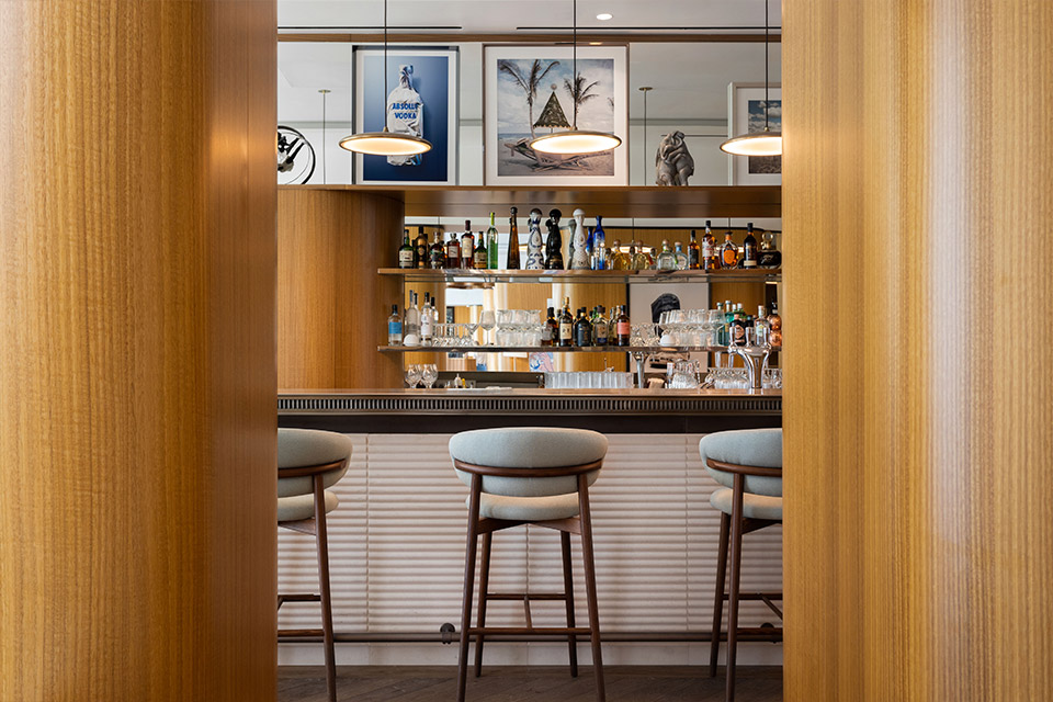 Bar with grey and wooden frame stools, shelves of liquor and glassware, pendant lights, and two wooden columns in the foreground