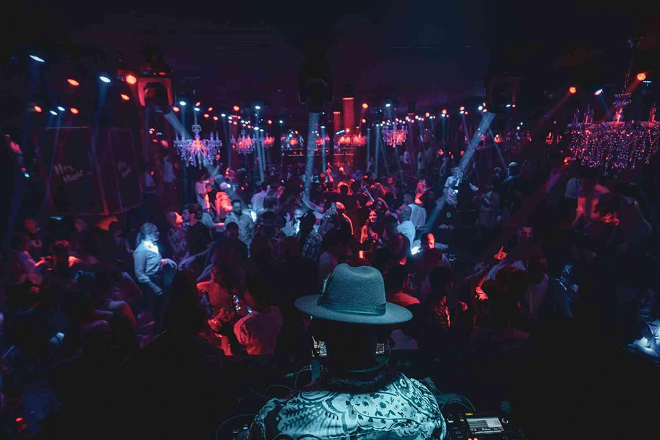 Danseurs dans une discothèque sombre aux lumières bleues et rouges avec un DJ coiffé d'un chapeau mou vu de dos.