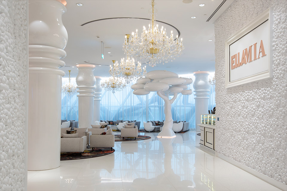 White dining room with four elegant glass and gold chandeliers, textured white wall with a pink sign that says EllaMia, and white block lounge chairs throughout, a white tree sculpture stands in the middle.