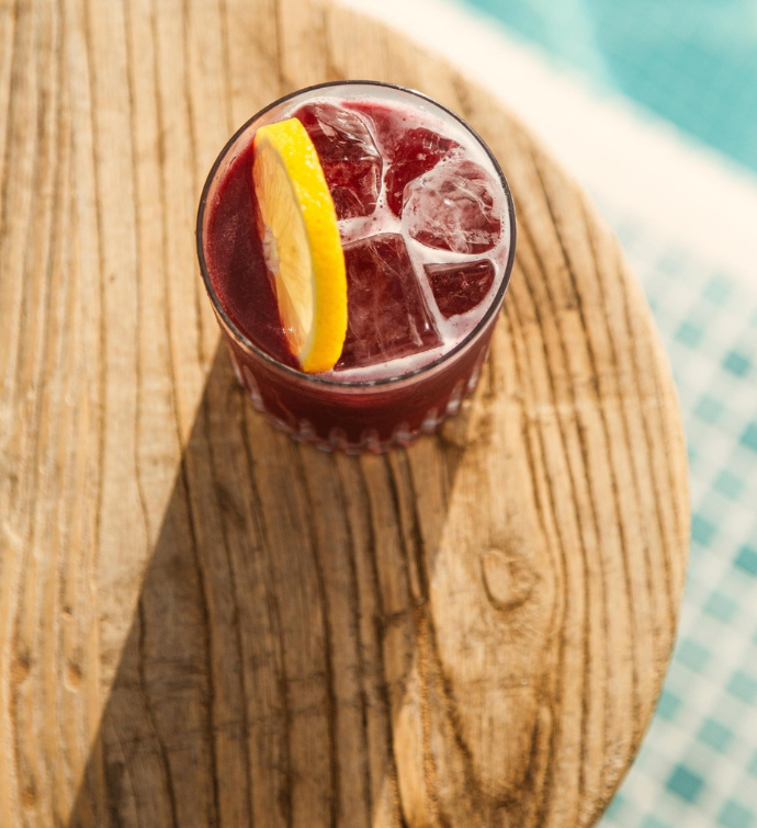 a purple cocktail garnished with a lemon on a wooden table with blue tiles in the background