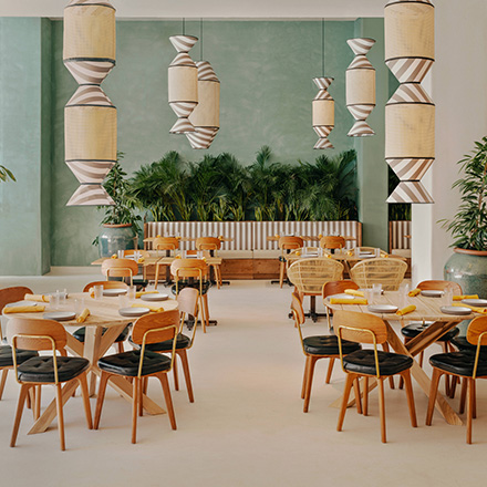 Comedor verde claro con lámparas colgantes a rayas, mesas redondas de madera con sillas de madera y palmeras al fondo.