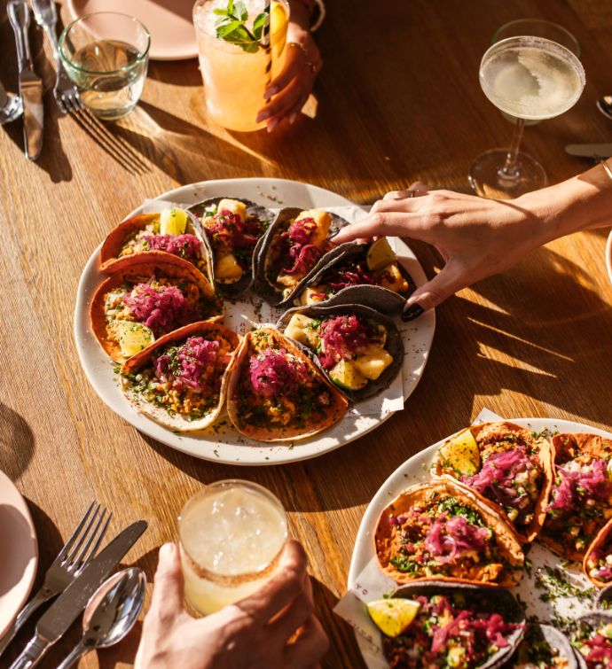 Two large plates of tacos on a light wooden table with hands holding cocktails