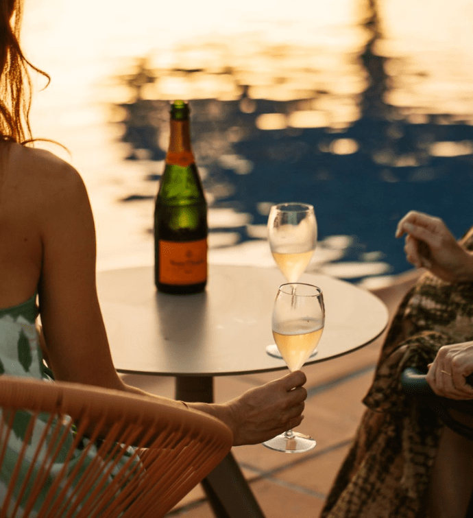 dos personas sentadas en una mesa con una botella de vino blanco mirando a la piscina durante la <em>golden hour</em>